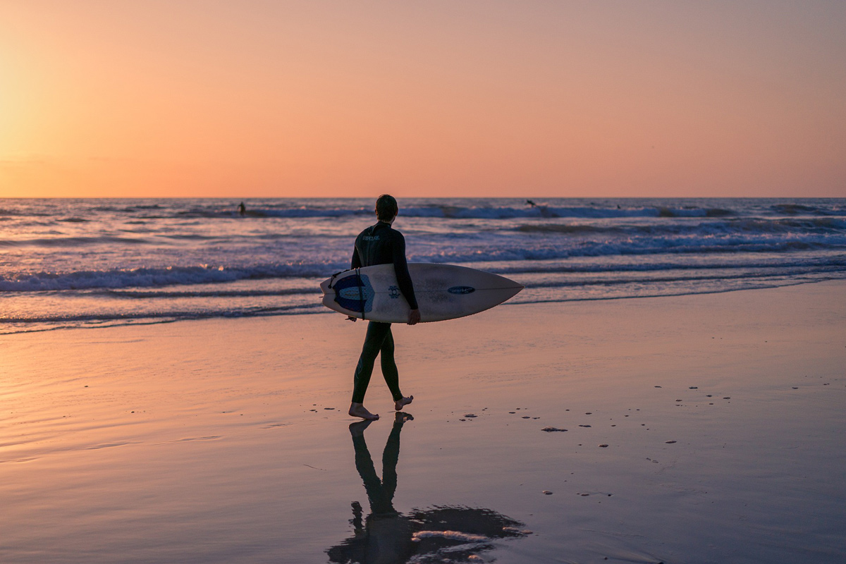 A surfer walks a San Diego beach at twilight
